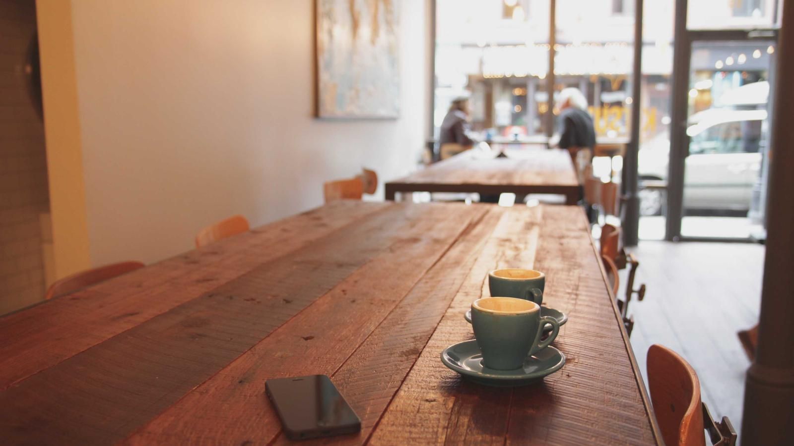 Een lange houten tafel in een licht café, met twee koppen koffie en mensen op de achtergrond